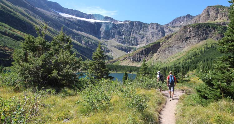 Two hikers walk towards a blue lake with trees and mountains all around.