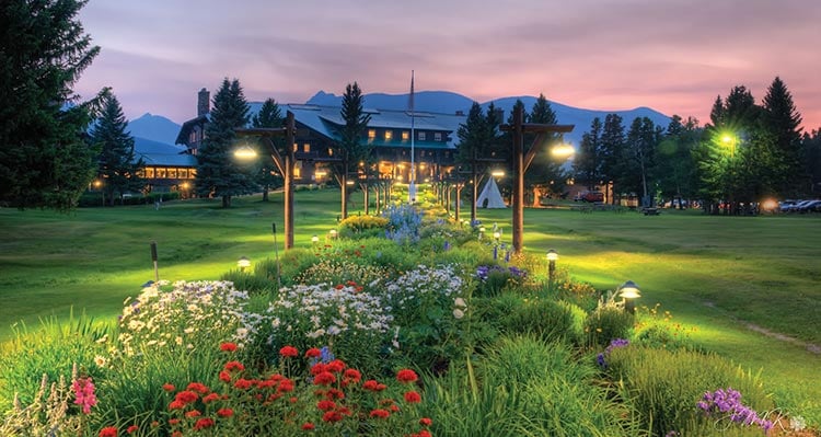 Flowerbed gardens stretch across a green lawn towards a large wooden lodge.