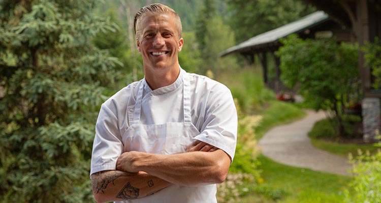 Chef Earl Reynolds stands in front trees and wooden cabins.