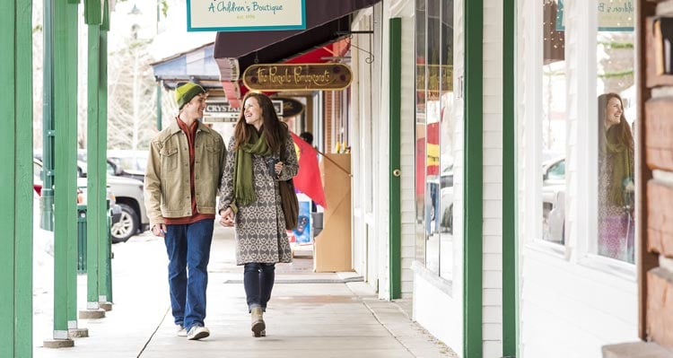 Two people walk along a street with shops and awnings.