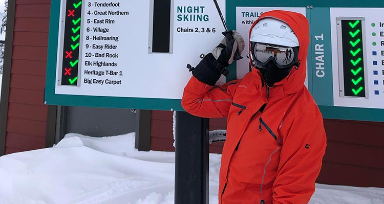 A skiier in an orange jacket next to an information board.