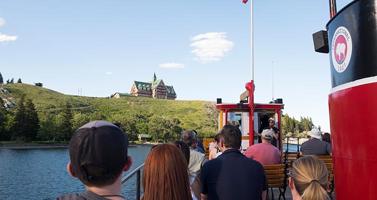 A group of people on a boat look up towards the Prince of Wales Hotel.