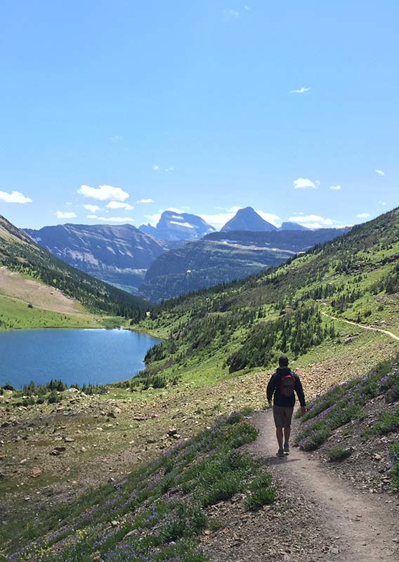 A hiker walks on a trail towards a small lake between tree-covered mountains.