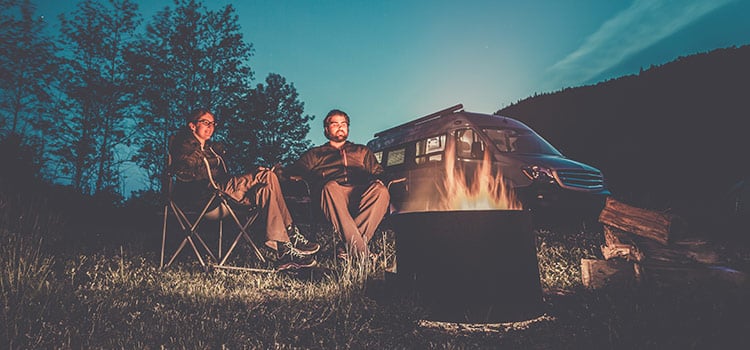 A couple sits by a campfire at dusk with a camper van in the background