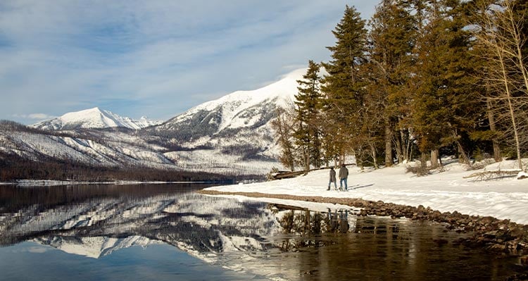 Two people snowshoe along a snowy lakeshore beside a conifer forest.