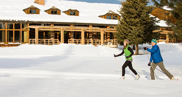 Two people cross-country ski along snowy trails next to a wooden hotel.