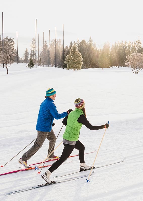 Two people cross country ski along a snowy meadow.