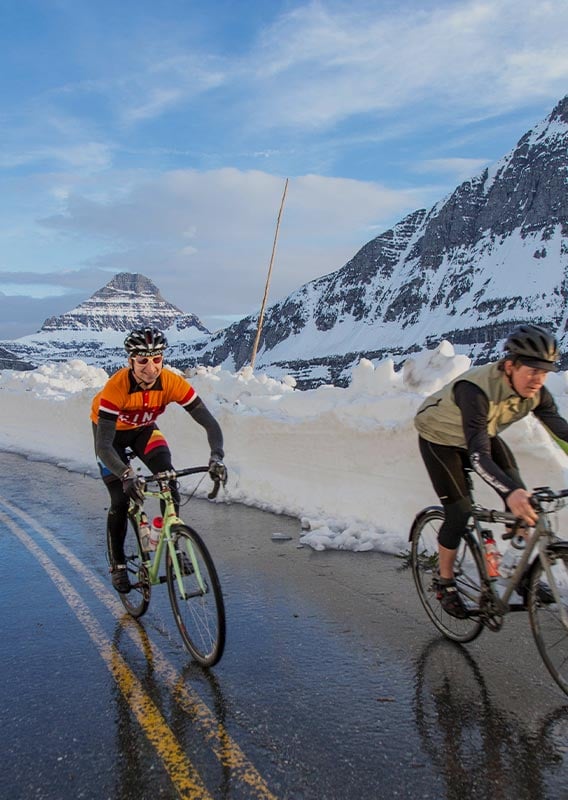 Two cyclists ride up the snowy road, mountains in the distance