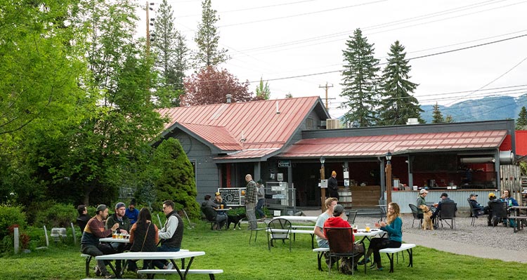 Groups of people sit at patios tables outside of a building, mountains in the background.