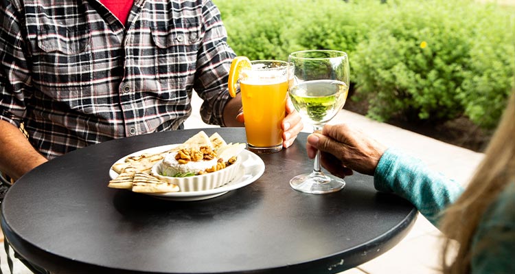 A couple sit at patio table with a beverage in hand and an appetizer on the table.