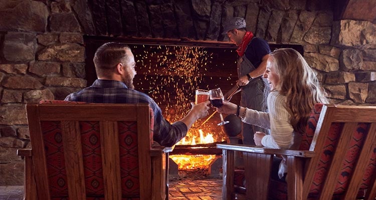 A couple sits in front of a large lit fire place enjoying a beverage. A man fueling the fire in front.