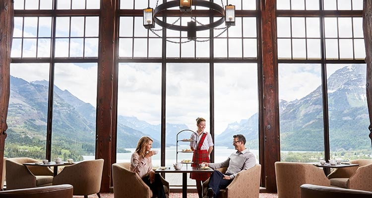A waitress serves a couple high tea in front of large window overlooking mountains and a lake.