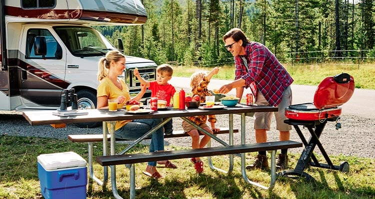 A family with young kids share a meal on a picnic table, an RV parked behind.