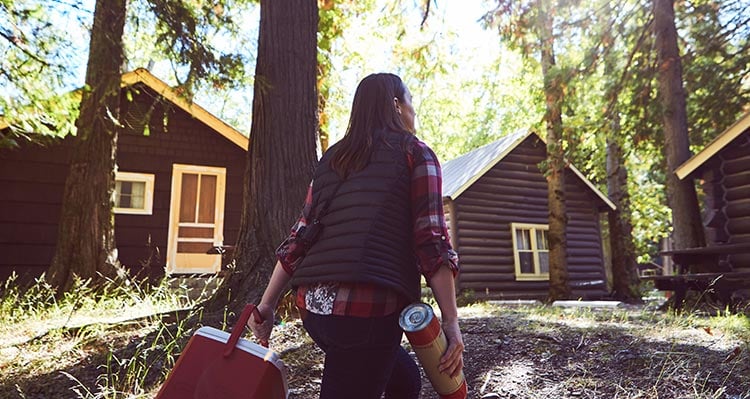 A woman walks toward cabins in the woods.