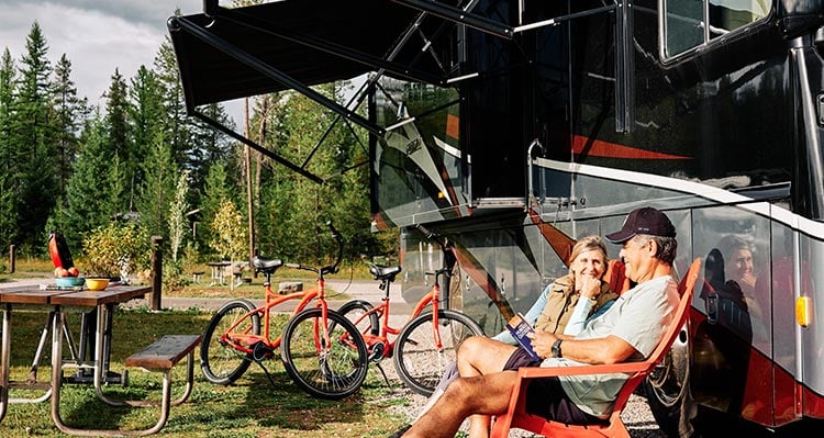 A couple sits outside of there RV enjoying the sun surrounded by forest.