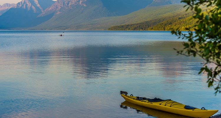 A yellow kayak sits at the shore of a still lake, mountains behind.