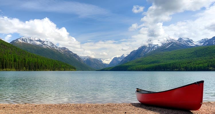 A red canoe sits at the shore of a lake, mountains and forest in the distance.