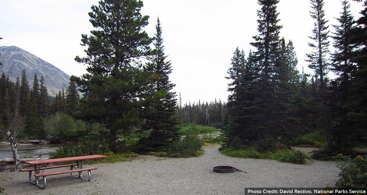 A picnic table and fire pit in a campsite surrounded by trees and a river.