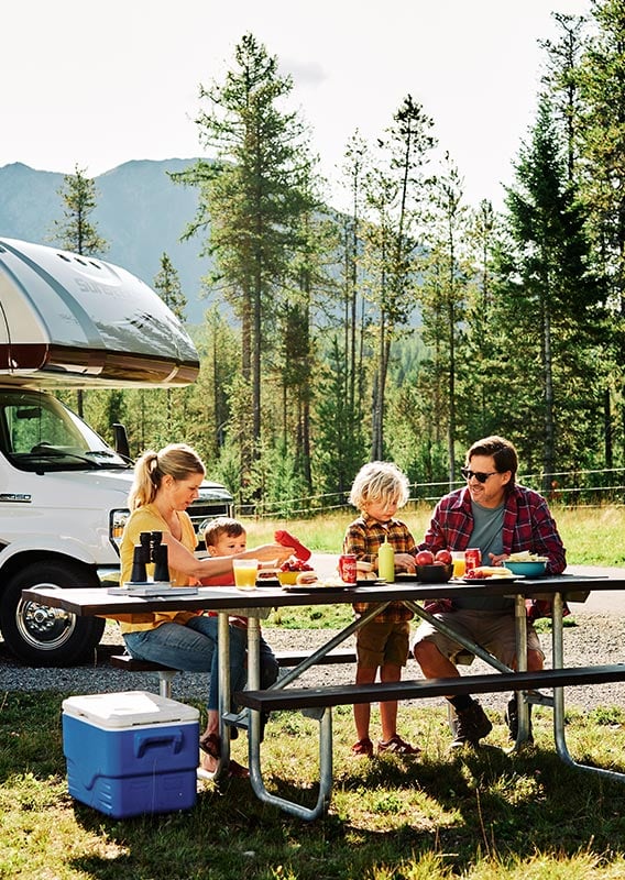A family sits at a picnic table having dinner, an RV behind.