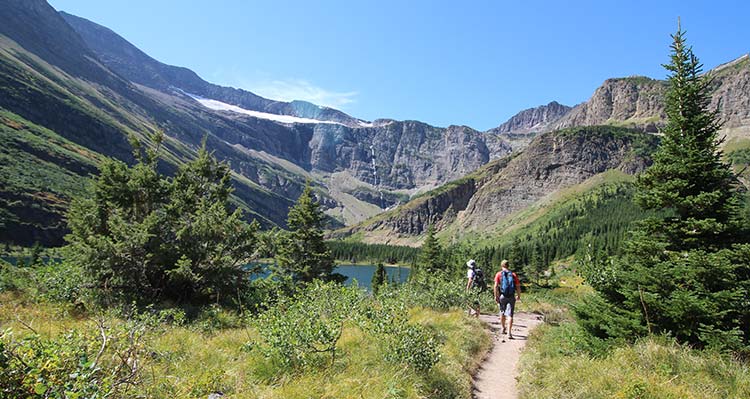 Two hikers in the distance walk toward a body of water, surrounded by mountains and trees in the summer.