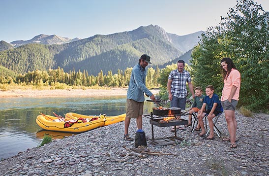 A river guide barbecues on a rocky river shore with a family.
