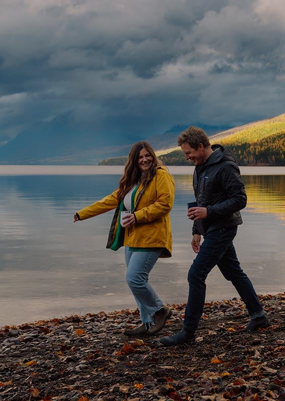 Two people walk on a rocky beach next to a large lake.