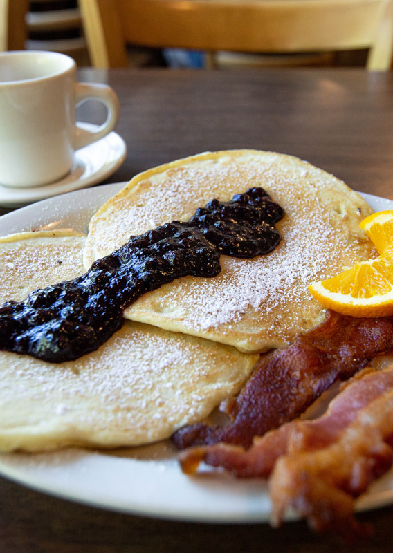 Pancakes with berry compote, bacon, and a slice of orange, with a cup of coffee in the background
