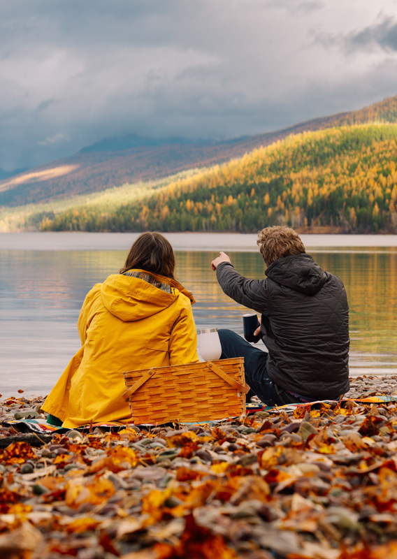 Two people sit on a colourful rocky beach by a large lake.
