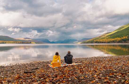 Two people sitting on a beach that is covered in fallen leaves, looking at the mountains and lake.