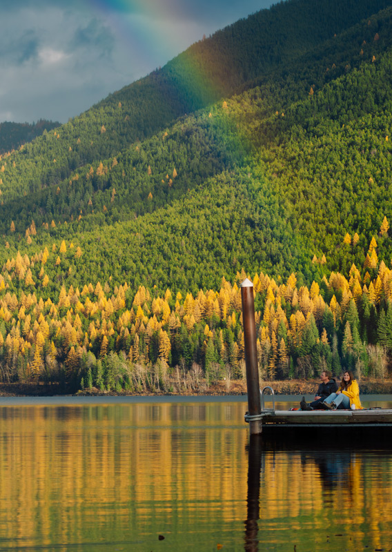 Two people sitting on a dock on a lake, looking at a rainbow, surrounded by fall colours.