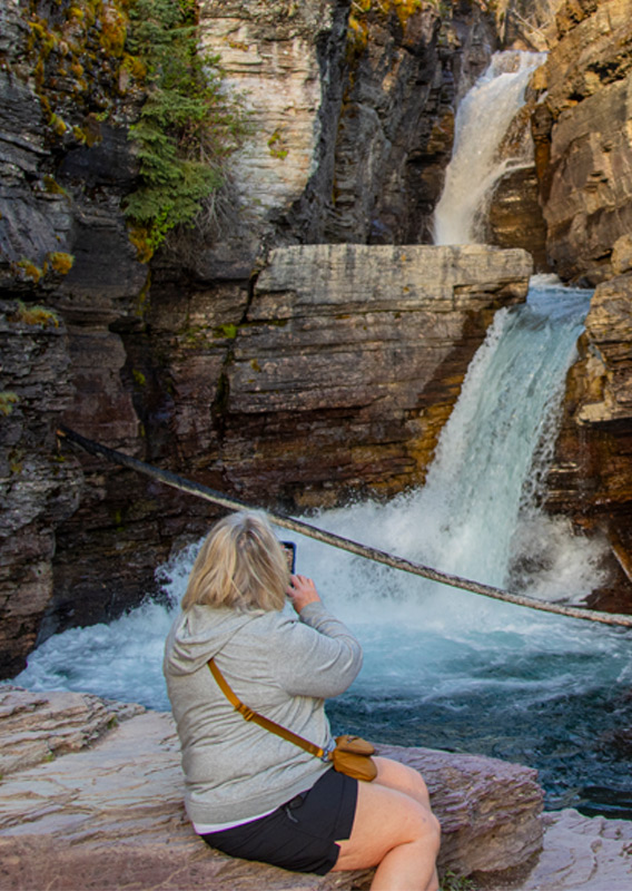 Two people sitting on rocks, taking a photo of a waterfall.