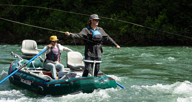 A guide rowing a boat along the Flathead River in West Glacier, Montana, while another guide stands on the boat fly fishing.