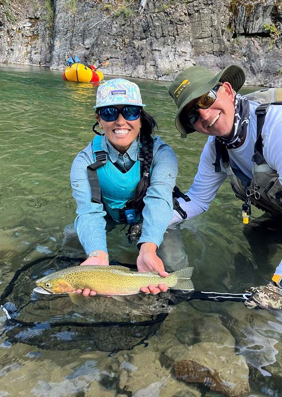 A fishing guide and a fisher hold a trout at the river surface.