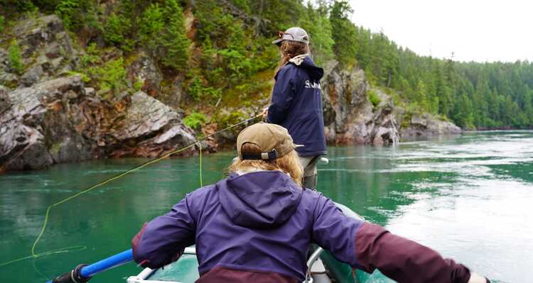 Two women in a fishing boat from behind while one rows and one stands.