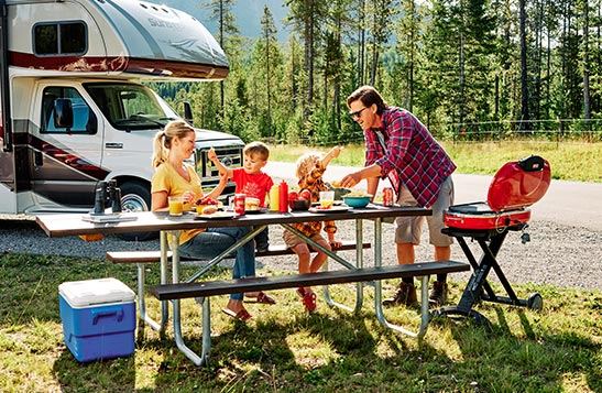 An older couple are sitting outside next to a firepit with their RV parked close at night.