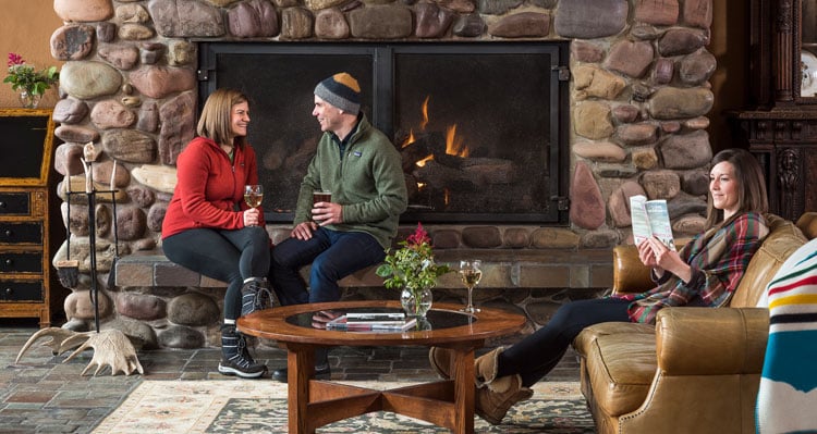 Two people sitting by the fireplace with drinks and one person reading in the lodge