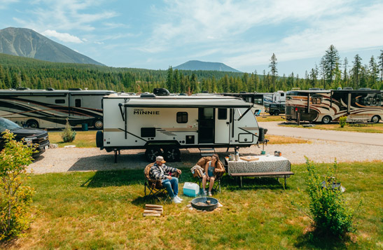 Two people sitting in camping chairs beside an RV and picnic table